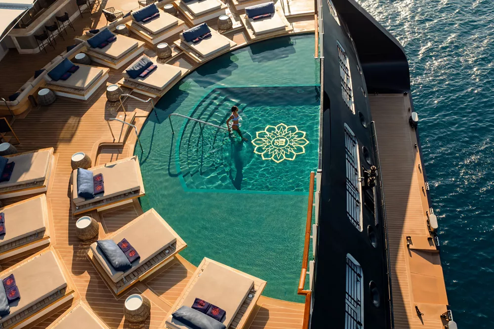 Luxury cruise ship pool deck with sun loungers and mosaic-tiled swimming pool overlooking the ocean.